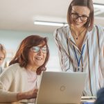 Professorin hilft einer Studierenden bei der Arbeit am Laptop. Im Raum sitzen weitere Studenten.